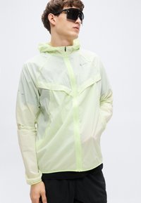 Ελαφρύ, διαφανές πράσινο μπουφάν με κουκούλα, κλείσιμο με φερμουάρ και ορατές ραφές. Διαθέτει αντανακλαστικές λεπτομέρειες και εμπρόσθιες τσέπες.