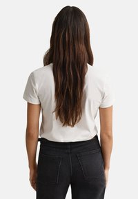 T-shirt branca de manga curta, vista de costas, combinada com jeans pretos de cintura alta. Cabelo castanho longo e ondulado parcialmente sobreposto à t-shirt.