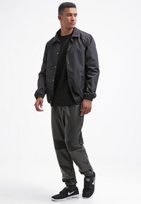 Czarna wodoodporna kurtka z zatrzaskami, noszona na teksturowanym czarnym swetrze. Szare tapered pants z siateczkowymi akcentami. Czarne sneakersy.