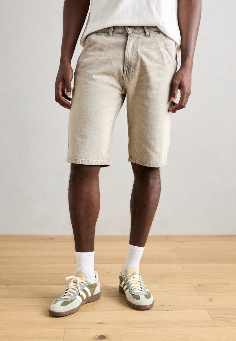 Beige denimshorts med en avslappnad passform, knappstängning och sidofickor. Kombineras med gröna och vita sneakers samt vita ankelsockor.