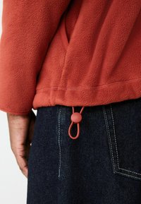 Roter Fleece-Pullover mit weicher Textur, ausgestattet mit einem cinched Saum, der durch einen roten Kordelstopper verstellbar ist. Kombiniert mit dunklem Denim-Stoff.