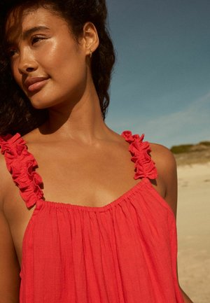 Robe sans manches rouge avec des bretelles volantes et un col froncé. Texture de tissu douce, présentant un design ample et fluide sur fond de plage.