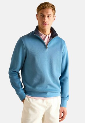 Light blue pullover sweatshirt with a zippered collar, textured fabric, and ribbed cuffs. Worn over a pink collared shirt.