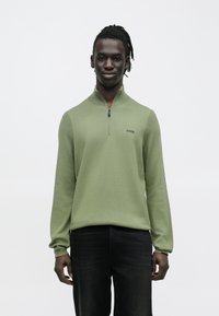 Zielony sweter na zamek z długimi rękawami, z prążkowanymi wstawkami i małym logo na piersi. Noszony z ciemnymi dżinsami, na neutralnym tle.