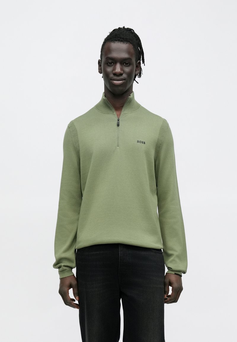 Zielony sweter na zamek z długimi rękawami, z prążkowanymi wstawkami i małym logo na piersi. Noszony z ciemnymi dżinsami, na neutralnym tle.