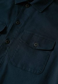 Ciemnoniebieska koszula z guzikami i kołnierzykiem, z dwiema kieszeniami na piersi i czarnymi guzikami; widoczna gładka faktura i złożenie materiału.