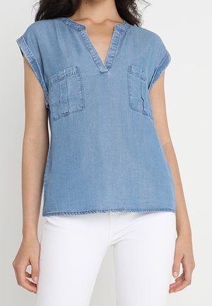 Woman wearing light blue sleeveless denim top with chest pockets and a V-neckline, paired with white pants.