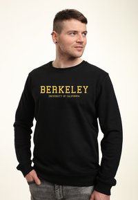 Felpa nera con maniche lunghe e scollo tondo, caratterizzata da un audace testo giallo: "UNIVERSITÀ DI BERKELEY CALIFORNIA." Texture morbida.