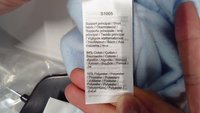 Vászon és poliészter ápolási címke világoskék szöveten. A címkén többnyelvű anyagösszetétel található: 84% pamut, 16% poliészter.