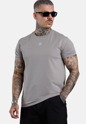 Man with short hair, sunglasses, and tattoos wearing a gray T-shirt and black pants, standing with one hand in pocket.