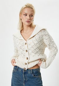 Strickjacke in Creme mit komplexen Mustern, gerippten Bündchen und Saum sowie großen braunen Knöpfen. Verfügt über einen breiten Kragen und einen strukturierten Stoff.