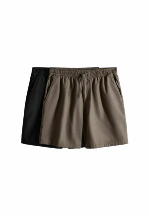 Bărbat purtând o cămașă maro cu nasturi și mâneci suflecate, pantaloni scurți asortați și pantofi maro slip-on, stând în fața unui fundal simplu deschis.