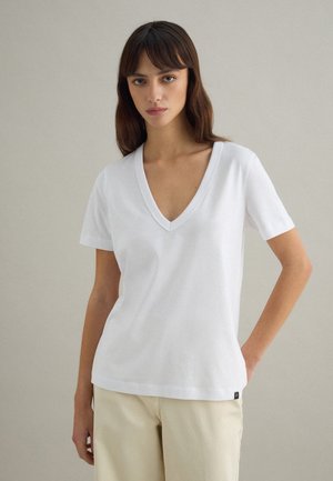 Frau mit braunen Haaren, die ein weißes V-Ausschnitt-Kurzarmshirt und beige Hose trägt, steht vor einem schlichten hellgrauen Hintergrund.