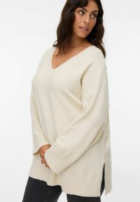 Sweter w kremowym kolorze z dzianiny, z dekoltem w serek, długimi rękawami i bocznymi rozcięciami. Miękka tekstura i luźny krój. W zestawie z ciemnymi dżinsami.