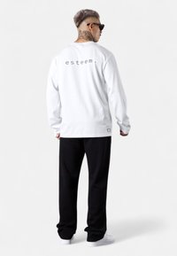 Weißes Langarmshirt mit dem Aufdruck "esteem" auf dem Rücken, kombiniert mit schwarzen Hosen und weißen Sneakers. Glatter Stoff, lockere Passform.