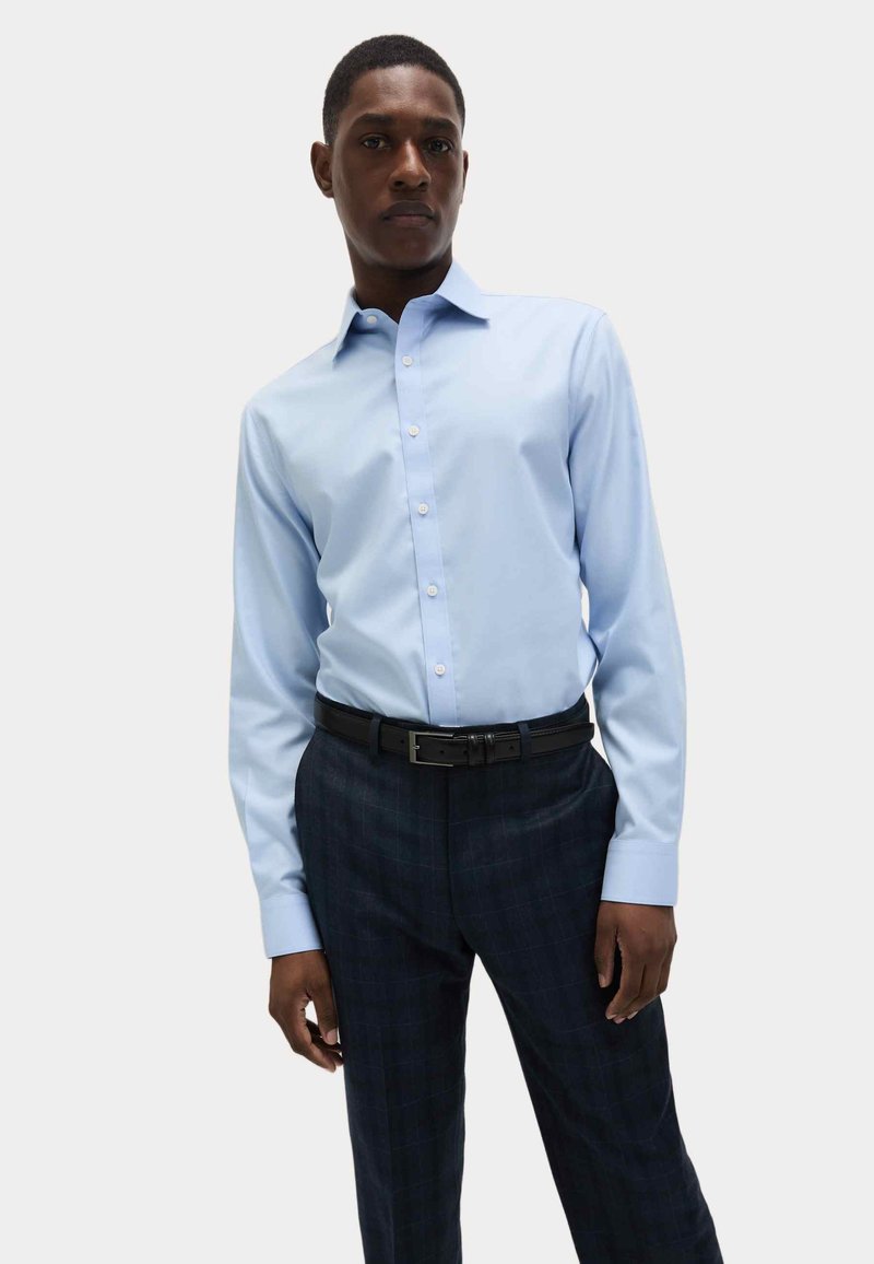 Lichtblauw overhemd met lange mouwen en een button-down kraag, gecombineerd met donker geruite broek en een zwarte leren riem.