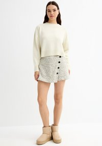 Suéter crema con dobladillo acanalado, acompañado de una falda gris con patrón y botones negros. Las botas beige hasta la rodilla completan el conjunto.