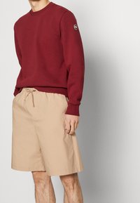 Felpa bordeaux con scollo rotondo e logo sulla manica, abbinata a pantaloni corti beige con cordoncino, realizzati in tessuto leggero e dotati di tasche laterali.