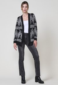 Cardigan nero con motivi geometrici bianchi, apertura frontale, maniche lunghe e dettagli frangiati sull'orlo, abbinato a jeans scuri e stivali neri.