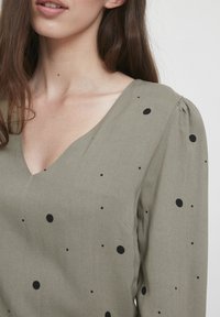 Olivgrüne Bluse mit V-Ausschnitt, die schwarze Punkte aufweist. Der Stoff hat eine glatte Textur und gepuffte Details an der Schulter.