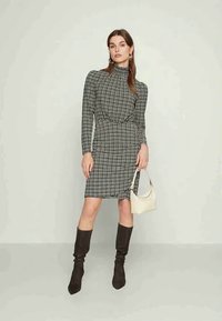 Futrované šaty s dlhým rukávom v čierno-bielych károvaných vzoroch s vysokým výstrihom, naťahovacím pásom a hladkou textúrou. Doplněné krémovou kabelkou a hnedými čižmami.