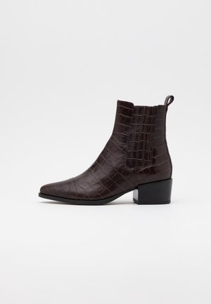 Mørkebrun ankelstøvle med krokodilletekstur, elastisk sidepanel, løkke bagpå, spids tå og klods hæl mod en hvid baggrund.
