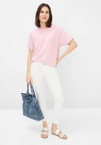 Lyserød kortærmet top, hvide slim-fit jeans og lyse sandaler. Holder en blå denim tote bag; afslappet outfit med løs pasform og minimalistisk design.