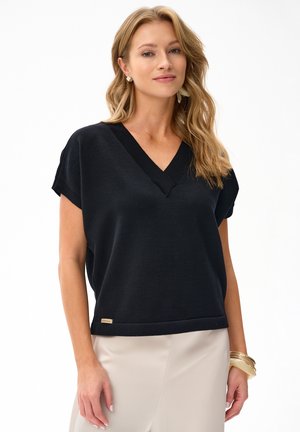 Blondynka ubrana w czarną bluzkę z krótkim rękawem i dekoltem w kształcie litery V oraz beżową spódnicę, z złotymi bransoletkami i kolczykami, stoi na białym tle.