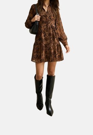 Frau trägt ein braunes, mit Leopardenmuster bedrucktes, geknöpftes Kleid, schwarze kniehohe Stiefel und trägt eine schwarze Umhängetasche.