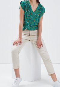 Grüner Blumenshirt mit V-Ausschnitt und kurzen Ärmeln, kombiniert mit beigefarbenen Hosen mit umgeschlagenem Saum und weißen Sneakers auf einem weißen Podest.