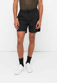 Black swim shorts with a drawstring waist, lightweight fabric, and a small logo accent on the left leg, paired with white sneakers and black socks.