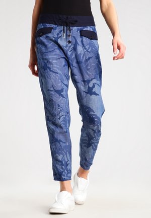 Pantalones con patrón de camuflaje azul, bolsillos con botones y cintura con cordón, usados con zapatos blancos sin cordones.