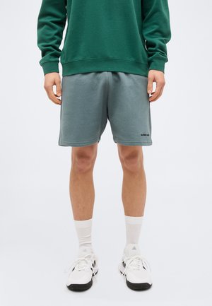 Άτομο που φοράει γκρι σορτς με μικρό κείμενο στο αριστερό πόδι, πράσινο φούτερ, λευκές κάλτσες κανονικού μήκους και λευκά αθλητικά παπούτσια με μαύρες σόλες.