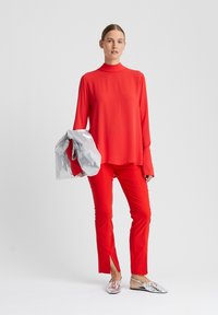 Rote Bluse mit langen Ärmeln und hohem Kragen, kombiniert mit passenden roten engen Hosen mit Schlitzen. Das Model hält eine silberne Jacke und trägt silberne Schuhe.