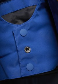 Blauer Stoff mit glatter Textur, der zwei Knöpfe aufweist: einen runden blauen Knopf und einen silbernen dekorativen Druckknopf.