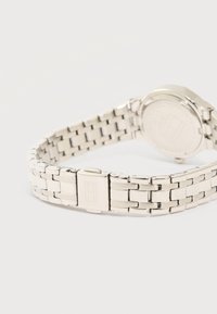 Ασημένιο μεταλλικό ρολόι χειρός με συνδεδεμένο βραχιόλι και κούμπωμα, τοποθετημένο ανάποδα σε λευκή επιφάνεια.