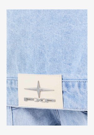 Lichtblauwe denimstof met een leren patch waarop een metalen zilveren sterrenlogo en de letters "PXP" zijn weergegeven. Zichtbare stiksels en textuurdetails.