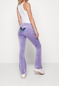 Lila sammet flarebyxor med fjärilsbroderi på bakfickan. Tyget har en jämn textur, kompletterad med en vit linne och vita sneakers.