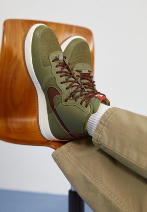 Personne portant des baskets vert olive et marron avec des semelles blanches, reposant les pieds sur une chaise en bois, vêtue d'un pantalon beige et de chaussettes blanches.