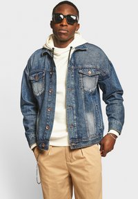 Denimjack in middelblauw met koperen knopen, twee borstzakken, relaxed fit. Draag het over een witte hoodie met trekkoorden. Lichtbeige broek.