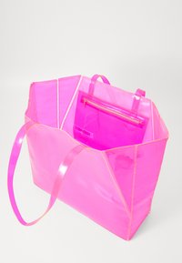 Transparente pinke Tragetasche mit kantigem Design, die eine kleinere pinke Tasche innen hat und gelbe Nähte betont. Zwei Griffe zum Tragen.