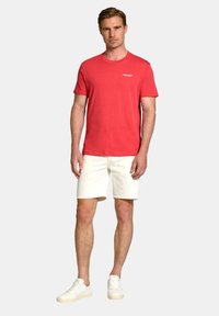 Tricou roșu din bumbac, cu guler rotund și mâneci scurte, asociat cu pantaloni scurți albi și adidași albi, stând în fața unui fundal neutru.
