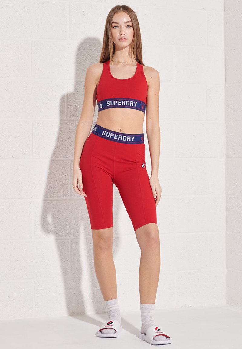 Rotes Sportoutfit bestehend aus einem ärmellosen Cropped-Top und hochgeschnittenen Shorts, beide mit marineblauen Akzenten und Branding. Kombiniert mit weißen Socken und Slides.