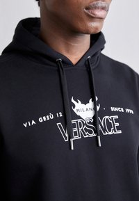 Melnā kokvilnas kapuce, ar baltu tekstu un grafikām. Iekļauta aukliņa kapuces un metāla galu. Dizaina elementi ir minimālistiski.