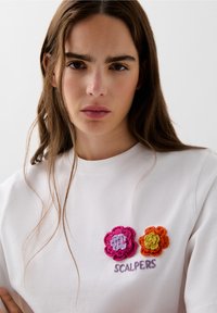 T-shirt bianco in cotone con due toppe floreali ricamate multicolori e la scritta "SCALPERS" in viola sotto.