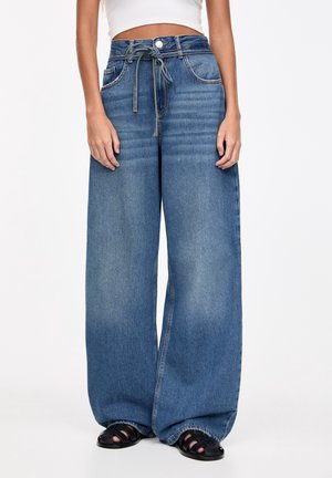 Femme portant un jean bleu taille haute à jambes larges avec une ceinture à cordon, un crop top blanc et des sandales noires à bout ouvert sur fond blanc.
