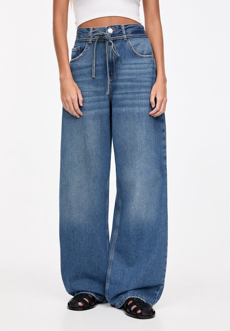 Femme portant un jean bleu taille haute à jambes larges avec une ceinture à cordon, un crop top blanc et des sandales noires à bout ouvert sur fond blanc.