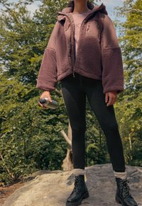 Osoba stojąca na zewnątrz na kamieniu, ubrana w różową polarową kurtkę, czarne legginsy, czarne buty, trzymająca w jednej ręce srebrną butelkę z wodą.