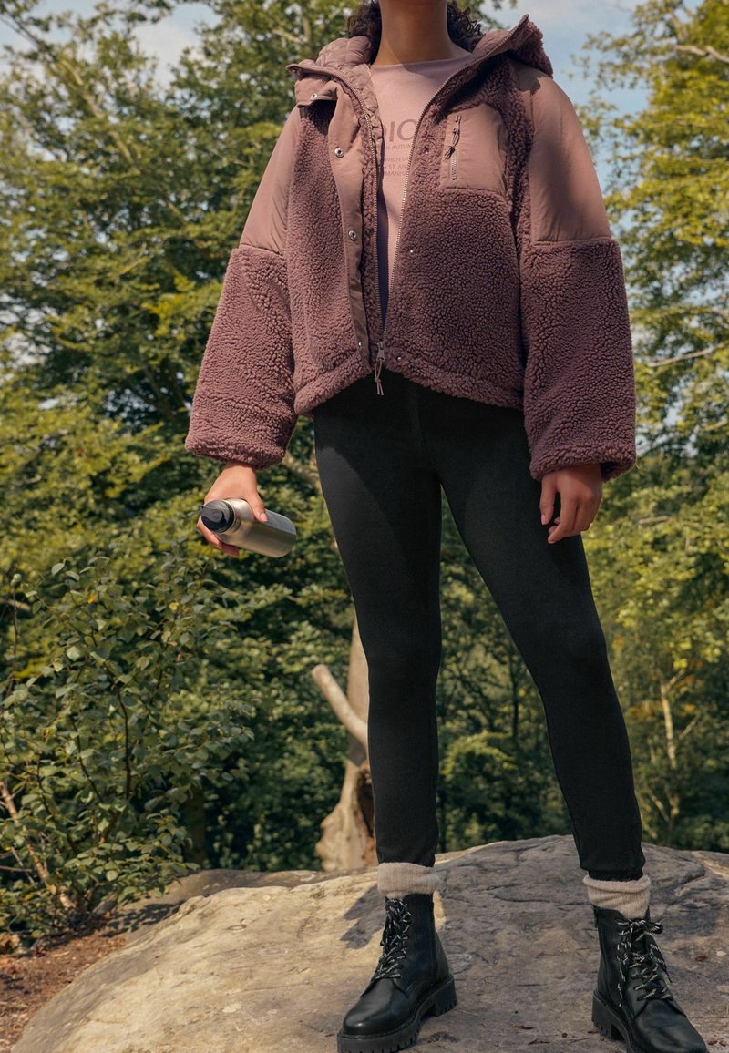 Osoba stojąca na zewnątrz na kamieniu, ubrana w różową polarową kurtkę, czarne legginsy, czarne buty, trzymająca w jednej ręce srebrną butelkę z wodą.