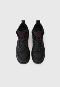 Černé turistické boty s texturovaným svrškem, polstrovaným límcem a kontrastními tkaničkami. Masivní gumová podrážka s drážkami pro lepší přilnavost. Detail červené značky.